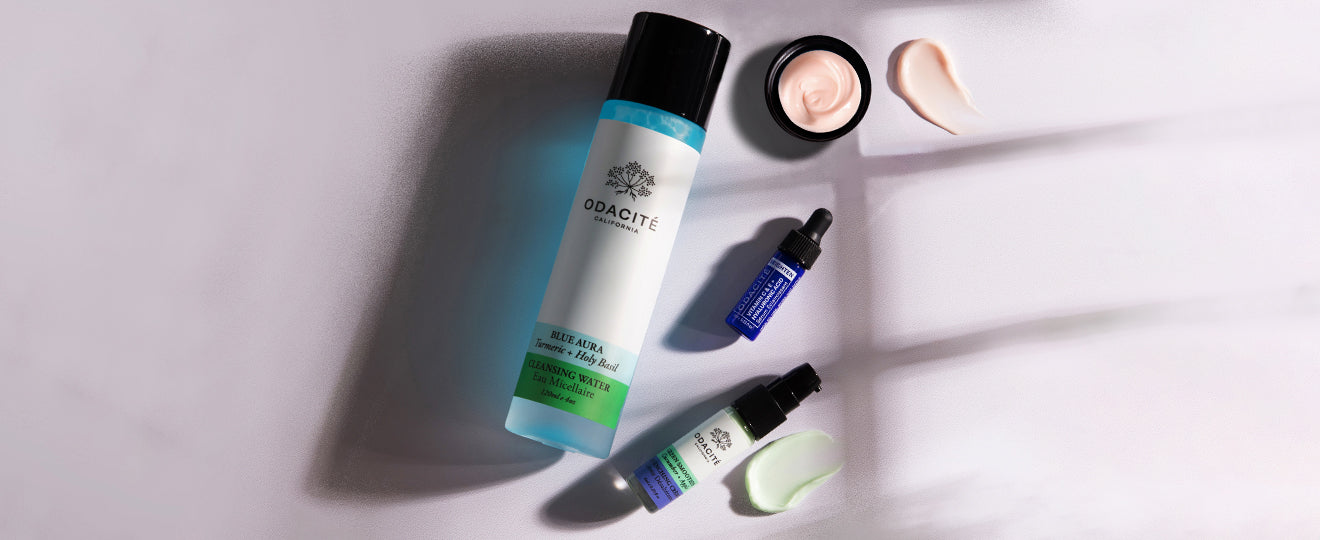 A bottle of Odacité cleanser, two blue dropper bottles, an open jar of cream, and swatches of cream and lotion are arranged on a light surface with soft shadows.