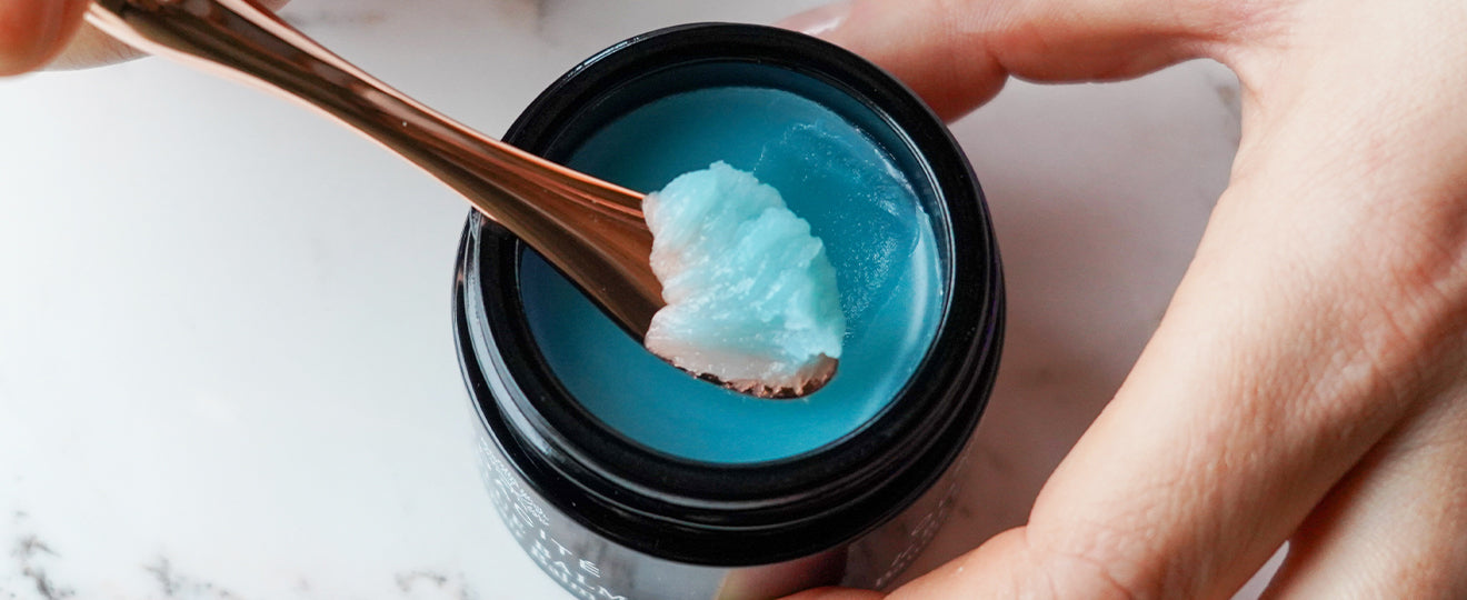 A hand holds a small jar of blue balm while a spoon scoops out some of the product, set against a white background.