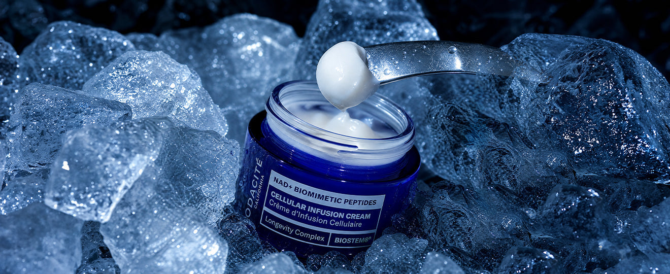 A jar of Odacité Hydra-Firm Body Cream sits open on a beige surface, with smooth, creamy white lotion spread around it.