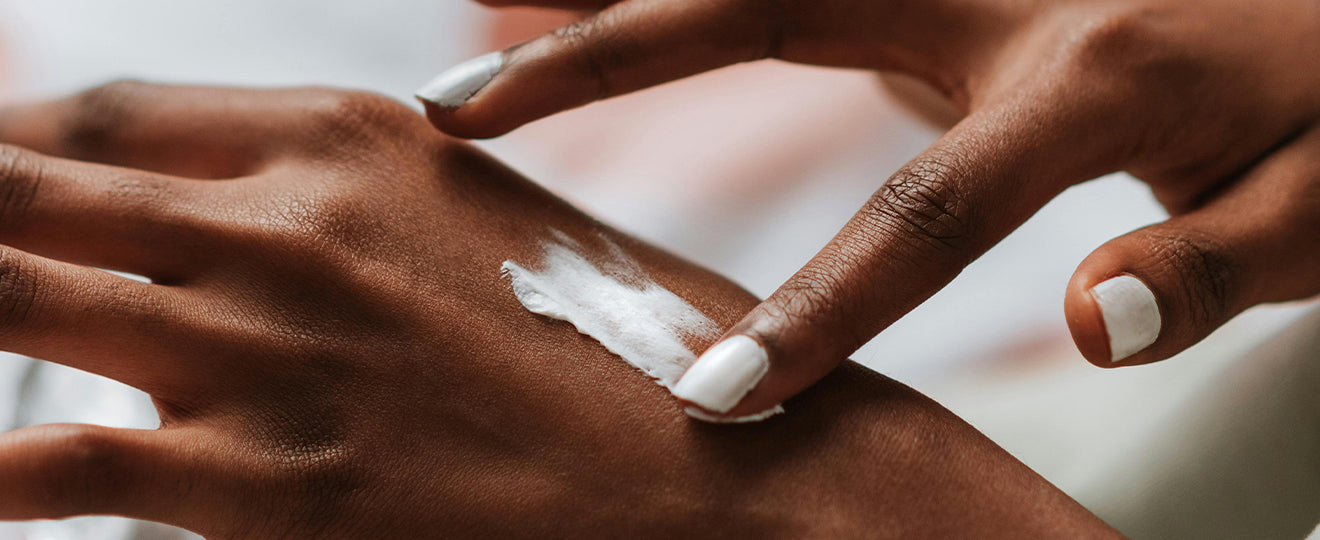 A person with light nail polish applies white lotion to the back of their hand, gently spreading it with their fingers.