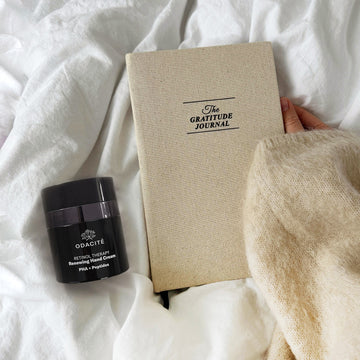 Hand holding a beige gratitude journal on a white bed next to Odacité Retinol Therapy Hand Cream with PHA + Peptides.