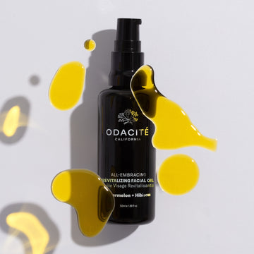 Odacite All-Embracing Watermelon + Hibiscus Facial Oil in a black bottle with yellow oil drops nearby.