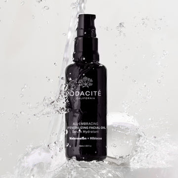Black bottle of Odacite All-Embracing Watermelon + Hibiscus Facial Oil with water splashes on a light background.
