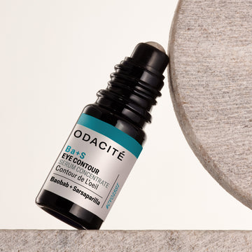 Odacite Ba+S Eye Contour Serum with rollerball in clear bottle, placed next to a textured stone surface.