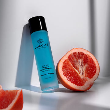 Odacité Blue Aura Turmeric + Holy Basil Micellar Cleansing Water bottle next to a grapefruit on a white surface.