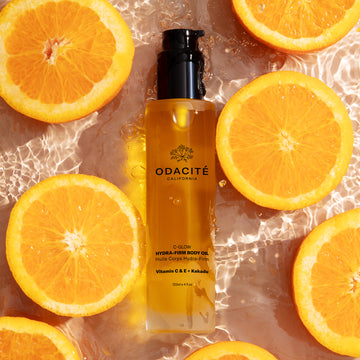 Odacité C-Glow Hydra-Firm Body Oil with Vitamin C & E + Kakadu next to orange slices on a wet surface.