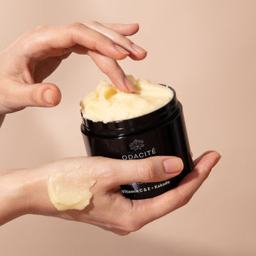 A hand scoops Odacité C-Smooth Hydra-Firm Body Polish with Vitamin C & E + Kakadu from a black jar and applies it to skin.