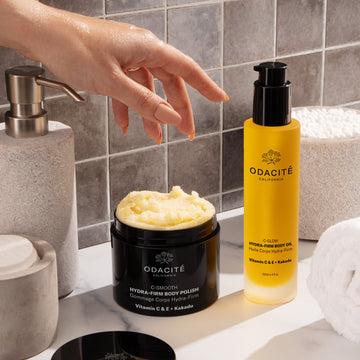 A hand scoops Odacité Hydra-Firm Full Body Duo, C-Glow & C-Smooth, from clear jars, showing creamy textures.