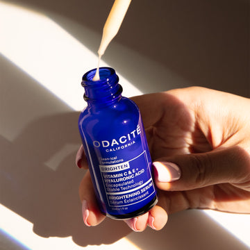 Hand holding Odacité Brightening Serum Vitamin C & E + Hyaluronic Acid; dropper dispenses serum above a blue bottle.
