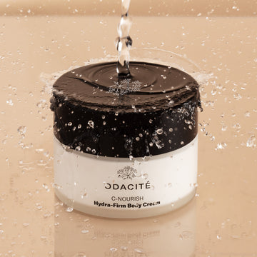 Odacité C-Nourish Hydra-Firm Body Cream jar with Vitamin C & E + Kakadu Plum splashed by water on a beige background.