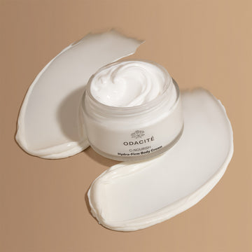 Odacité C-Nourish Hydra-Firm Body Cream, creamy white lotion swirled on a smooth beige surface.