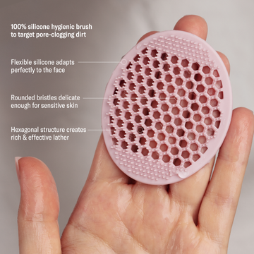 A hand holds the Odacité Pore & Face Cleansing Brush, a pink silicone brush with textured bristles.