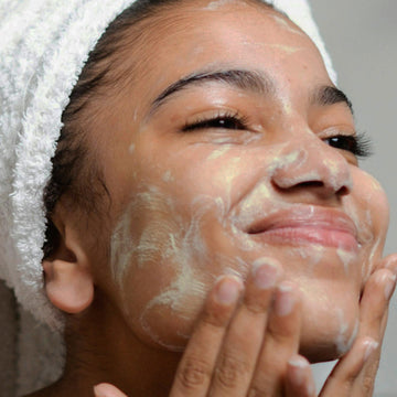 Person with a towel wrapped on their head uses Odacité Daily Calm Cleansing Duo to remove makeup from their face.