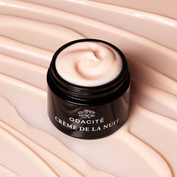 Open jar of Odacite Crème de la Nuit Vitamin C + Ashwagandha Night Cream on a swirled beige background; creamy texture visible.