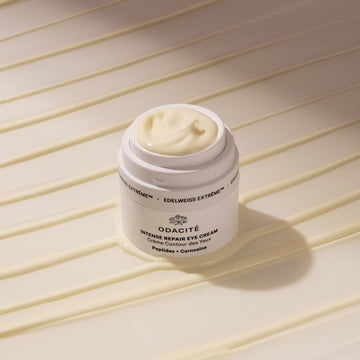 Open jar of Odacité Intense Repair Eye Cream Edelweiss Extrême + Peptides & Carnosine on striped background, cream visible inside.