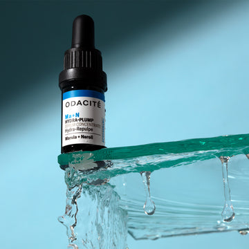 Odacite Hydra-Plump • Marula + Neroli • Ma+N Serum Concentrate dropper bottle on melting ice with water drops, blue background.