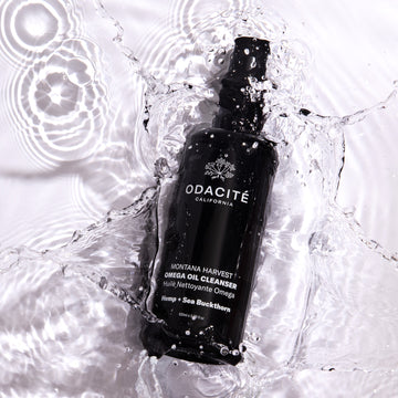 Black bottle of Odacite Montana Harvest Hemp + Sea Buckthorn Omega Oil Cleanser splashing in clear water, forming ripples.