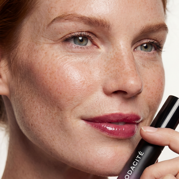 Freckled woman with glossy lips holds Odacité SPF 40 Nourishing Lip Serum tube near her chin.