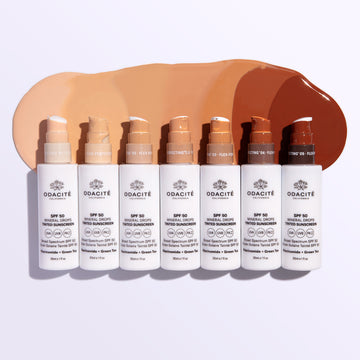 Seven Odacité SPF 50 Tinted Mineral Drops bottles in different shades, each with a matching swatch showing coverage.