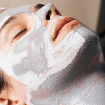 Person with eyes closed wears Odacite Synergie[4] 4-IN-1 Powder Masque, a light gray and white mask covering the face.