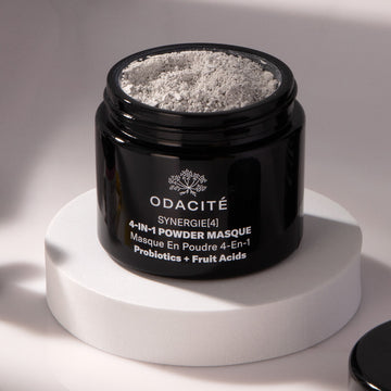 Odacite Synergie[4] Probiotics + Fruit Acids 4-in-1 Powder Masque in a black jar, gray powder visible, on a white platform.