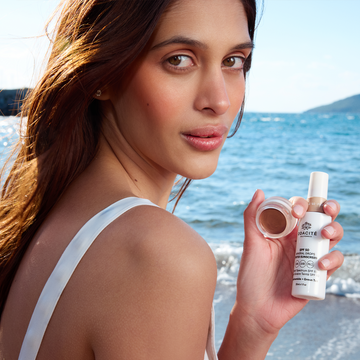 A woman at the beach smiles while holding Odacité SPF 50 Tinted Sunscreen & Color Balm Duo.
