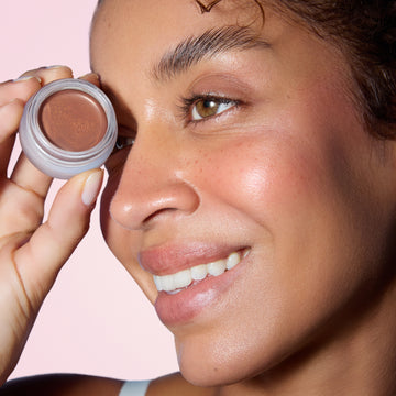 Woman smiles, holding Odacité SPF 50 Color Balm & Brush Duo near her eye; glowing skin, pink background.