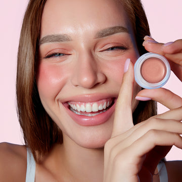 Woman smiles, holding Odacité SPF 50 Color Balm & Brush Duo close to her face with a light pink background behind her.