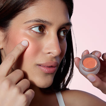 Woman applies Odacité’s SPF 50 Color Balm & Brush Duo in peach shade to her face with a finger, product in hand.