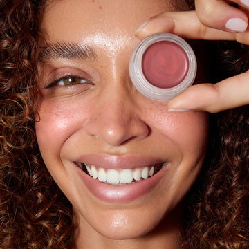 Woman with glowing skin smiles, holding an Odacité SPF 50 Color Balm & Brush Duo jar over one eye.