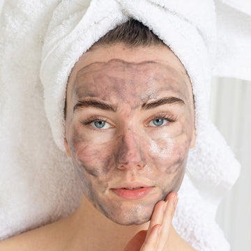 Woman with a towel on her head applies Odacite Black Mint • Charcoal + Rhassoul Clarifying Cleanser to her face.