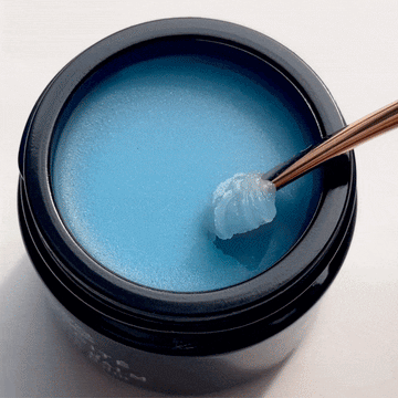 A spatula lifts Odacite le Blue Balm • Blue Chamomile + Shea Butter from an open black jar on a white background.