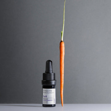 Odacite Smooth + Glow Mature Skin Duo serum bottle upright beside a whole carrot on a plain surface.