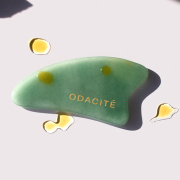 Odacité Crystal Contour Gua Sha Green Aventurine with gold lettering on a light surface, next to drops of yellowish oil.