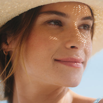Woman in a straw hat smiles outdoors under a blue sky, her skin highlighted while using Odacite's Tone + Glow Mature Skin Duo.