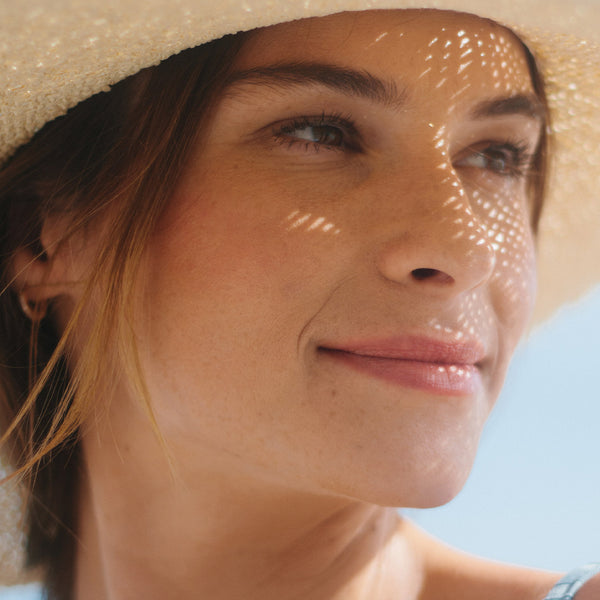 Woman in a straw hat smiles outdoors under a blue sky, her skin highlighted while using Odacite's Tone + Glow Mature Skin Duo.