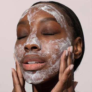 A woman applies Odacité Jojoba Pearls + Rooibos Tea Daily Hydra-Exfoliant, creating a white foamy lather on her face.