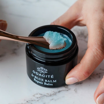 A hand uses a spatula to scoop Odacite le Blue Balm • Blue Chamomile + Shea Butter Repair Balm from a black jar on marble.