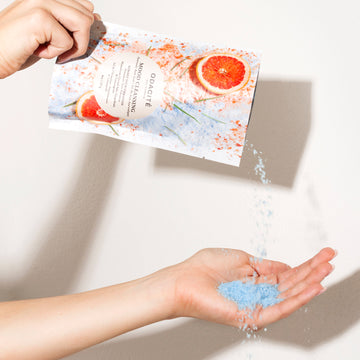 A hand pours Odacite Relax & Recenter Bath Set soak into its palm from a grapefruit-patterned packet against a white background.