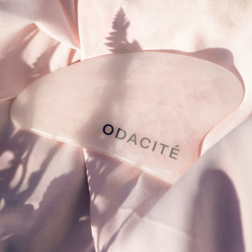 Odacite Crystal Contour Rose Quartz Gua Sha on blush pink fabric, with soft plant shadows in the background.