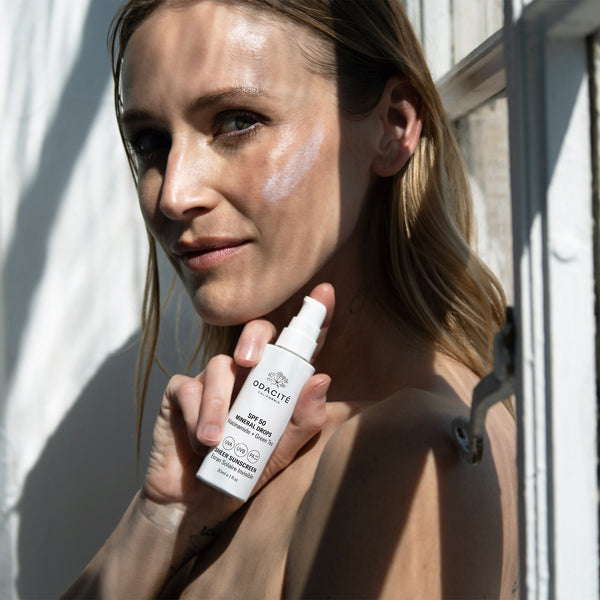 Woman applies Odacité Mineral Drops • Niacinamide + Green Tea SPF 50 Sheer Sunscreen by a sunny window.
