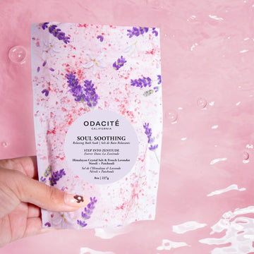 A hand holds Odacite's Soul Soothing Bath Soak with Himalayan Crystal Salt, French Lavender, Neroli & Patchouli over water.