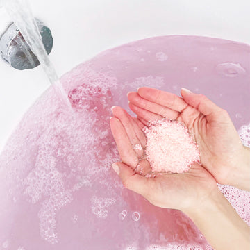 Hands holding Odacite Soul Soothing Bath Soak: Himalayan Crystal Salt, French Lavender Neroli + Patchouli above a pink-filled bath.